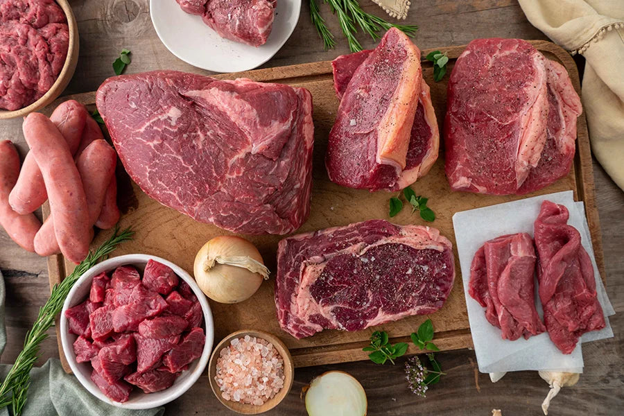 Various raw beef cuts styled on kitchen bench top, with herbs, vegetables and salt