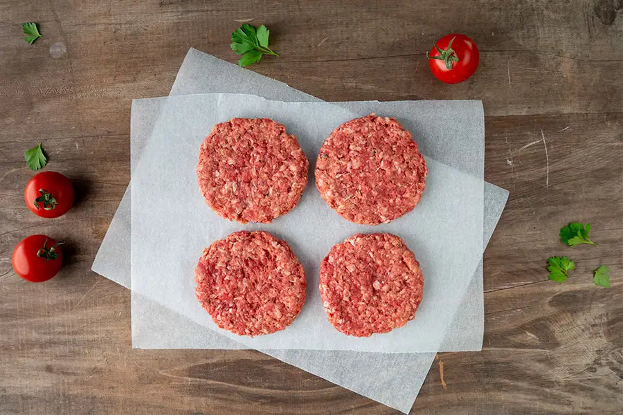 Four meat patties on a kitchen bench with herbs and tomatoes