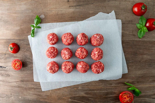 12 meatballs arranged on baking paper on a wooden kitchen top. Sliced tomato and herbs surrounding them