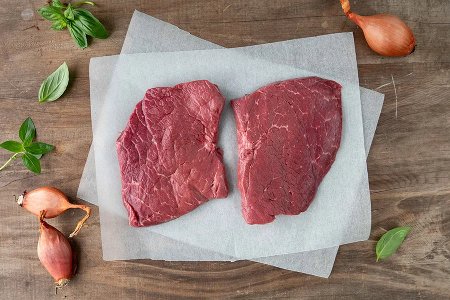 two topside steaks on benchtop with shallots and herbs